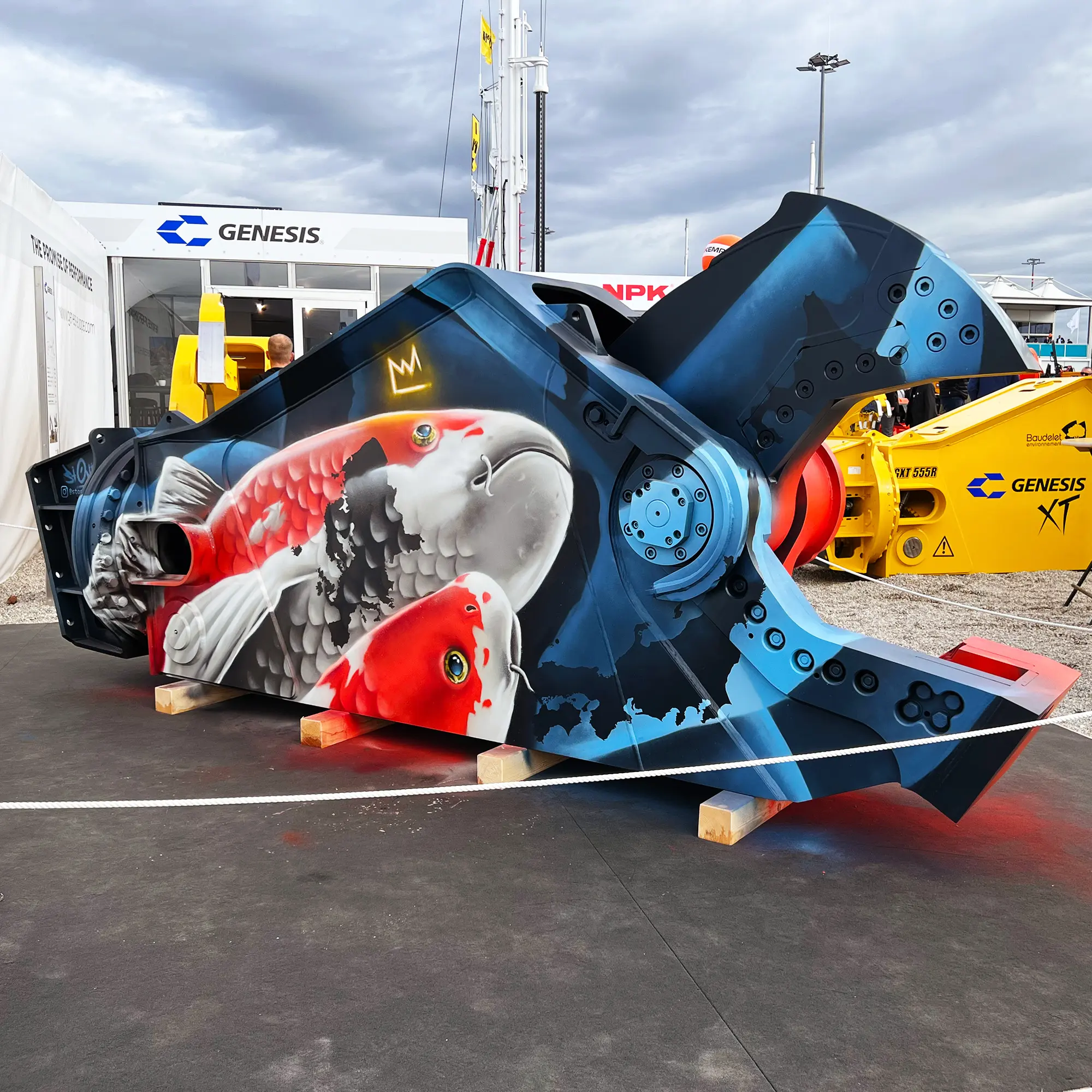 Großflächiges Graffiti auf einem industriellen Maschinenbauteil zeigt zwei realistisch gemalte Koi-Fische in Rot-, Weiß- und Schwarztönen, eingebettet in eine technisch anmutende, blau-schwarze Hintergrundgestaltung. Das detailreiche Graffiti verbindet organische Tiermotive mit mechanischen Formen und Schraubenelementen und ist auf einer massiven Metallkonstruktion im Außenbereich einer Messe oder Ausstellung angebracht.