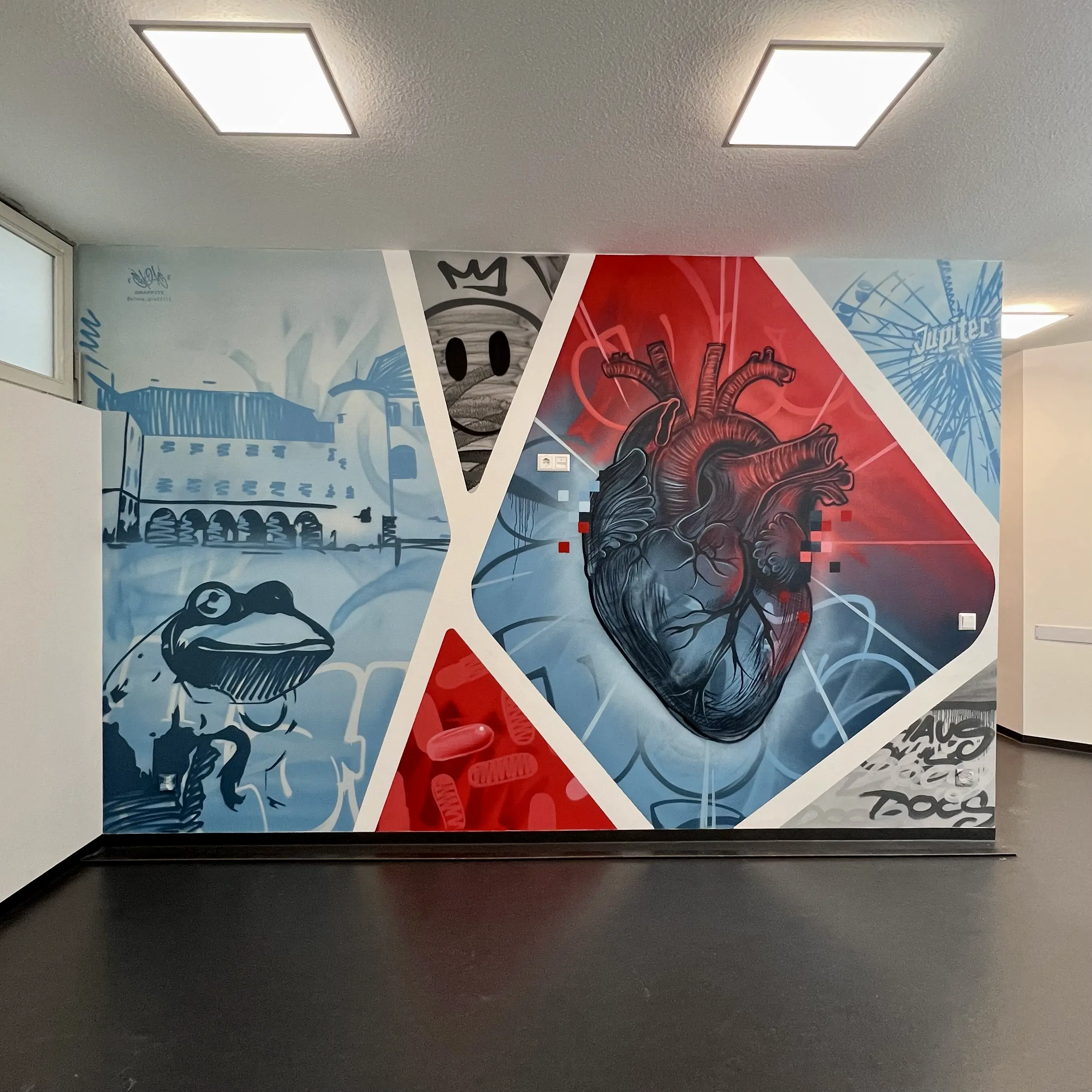 Großflächiges Graffiti an einer Innenwand zeigt eine collageartige Komposition aus verschiedenen Motiven, darunter ein anatomisch dargestelltes menschliches Herz in Rot- und Blautönen, abstrahierte Figuren, Schriftzüge und grafische Elemente. Das Wandgraffiti ist in geometrische Flächen unterteilt und verbindet realistische und illustrative Stile, wodurch ein moderner, künstlerischer Raumeindruck entsteht.